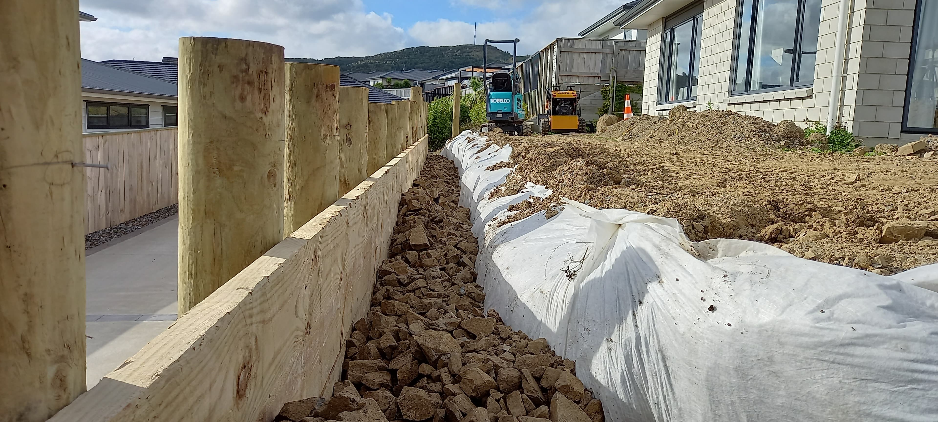 Timber pole retaining wall under construction with a gravel drainage trench and geotextile-lined bank in Wellington
